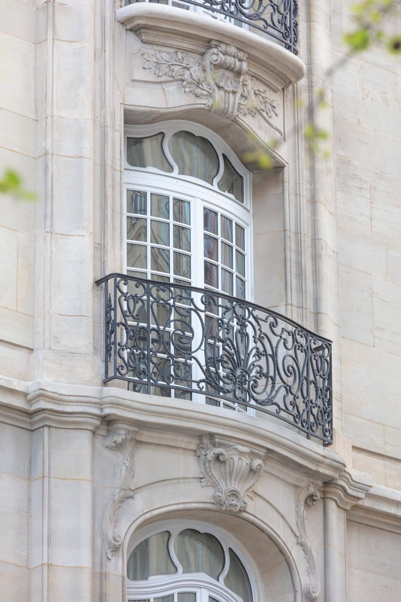 Fenêtre bombée à petits-bois sur une façade en pierre de taille sculptée, balcon en fer forgé, immeuble parisien